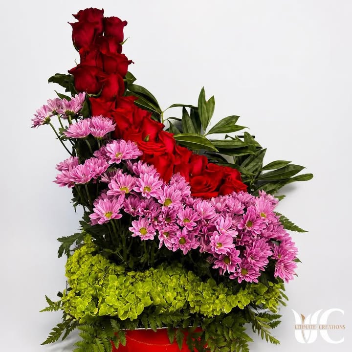 Red roses and pink flowers layered over green blooms and foliage in a red container