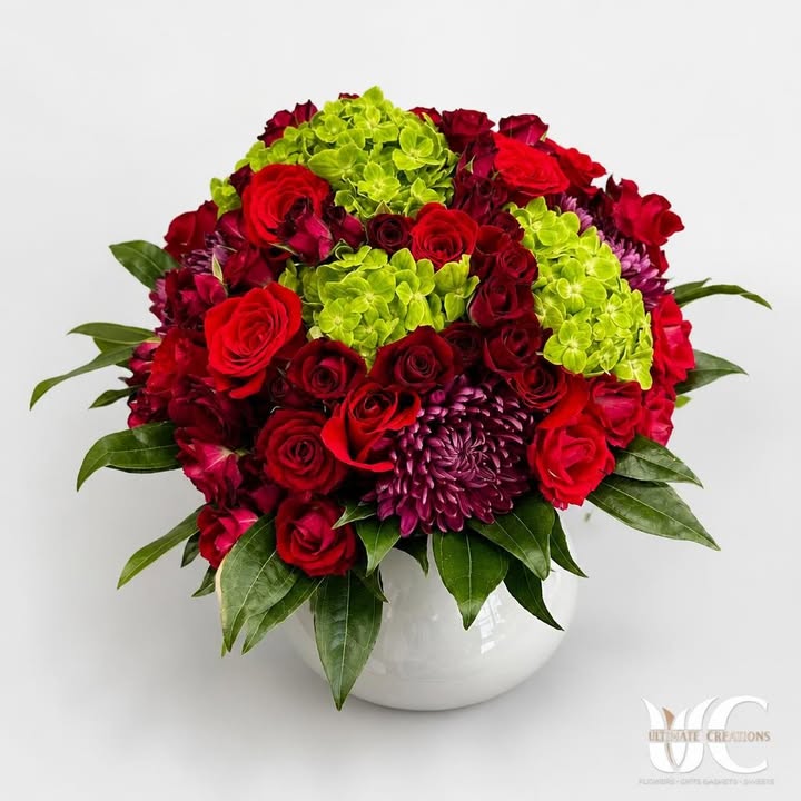 Rounded red and burgundy rose arrangement with green blooms, a purple focal flower, and glossy greenery in a white vase.