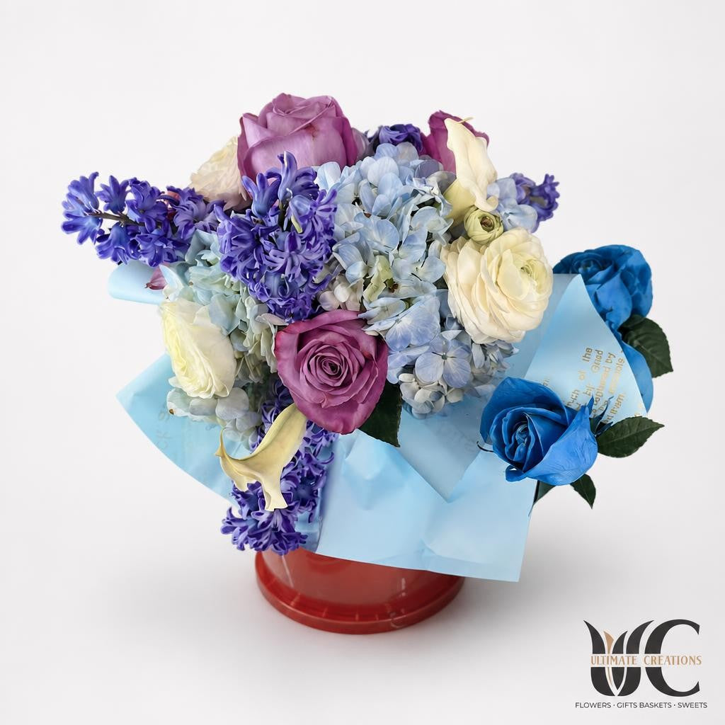 Lush multicolor bouquet in light blue wrapping, featuring pale blue clustered blooms, violet flowers, pink and coral roses, cream flowers, and a vivid blue rose.