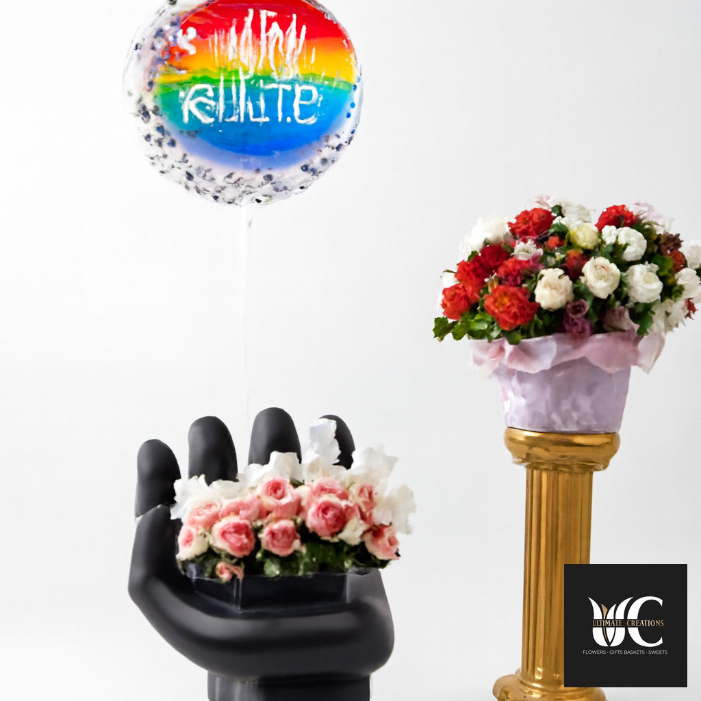 Rainbow-themed balloon above two floral arrangements, one in a black hand-shaped holder and one on a gold pedestal.