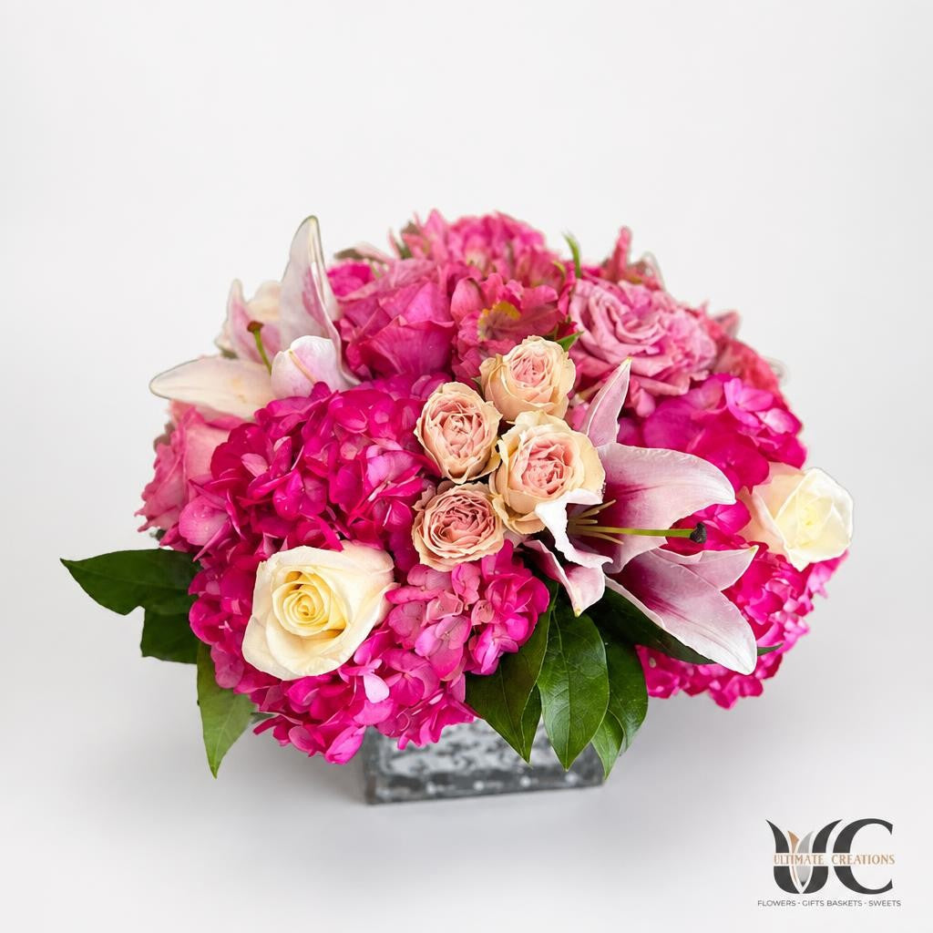 Rounded floral arrangement with vivid fuchsia blooms, cream and blush roses, pale pink lilies, and green leaves in a clear glass vase on a light tabletop