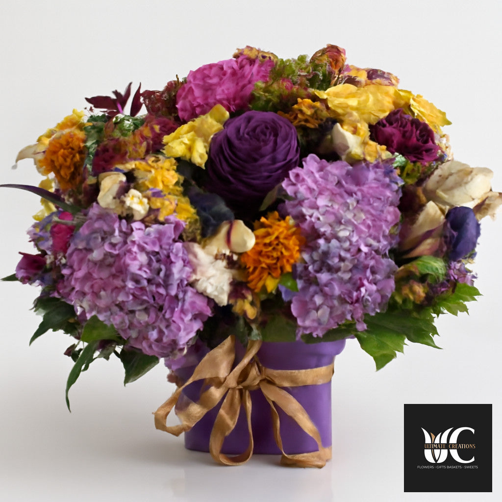 Purple flower box arrangement with mixed purple, lavender, yellow, and cream blooms tied with a gold ribbon.