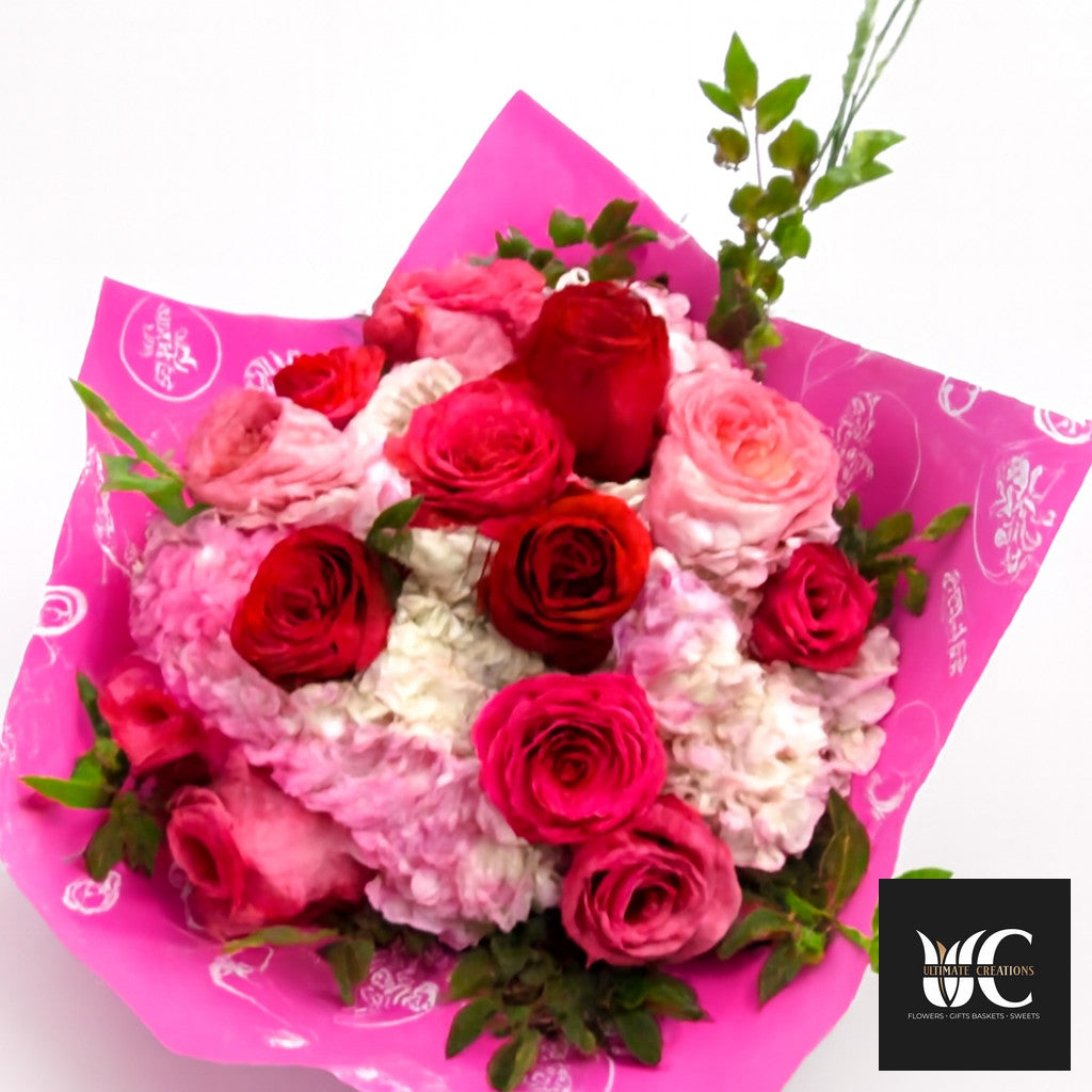 Hand-tied bouquet of pink and red roses with white flowers and greenery in bright pink wrapping paper.