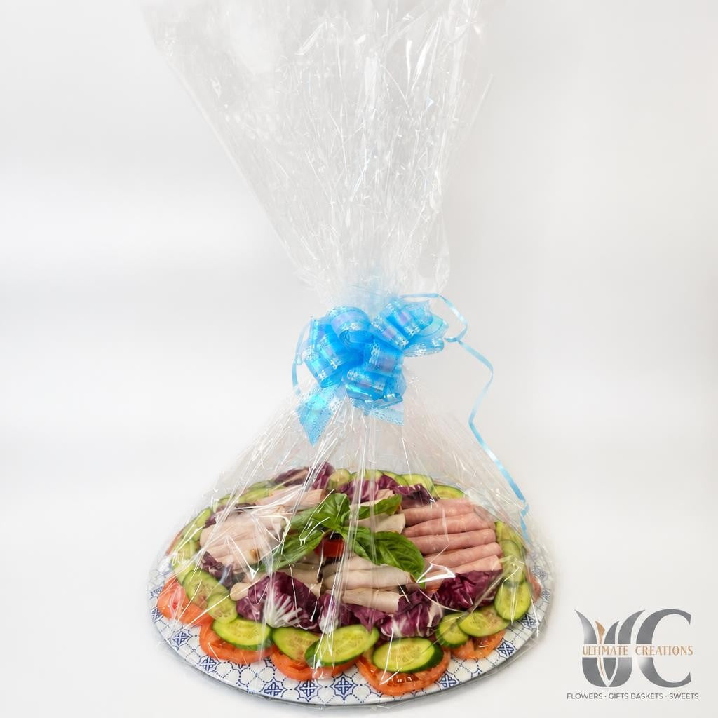 Cellophane-wrapped edible platter on a patterned plate, filled with sliced vegetables, leafy greens, and deli-style meats, tied with a bright blue bow.