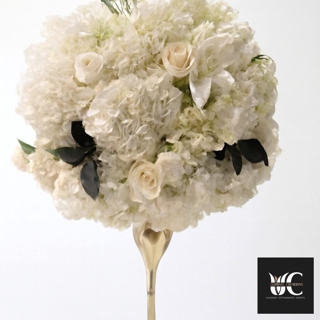 Ivory and cream floral centerpiece on a gold pedestal with soft rose accents and dark leaves.