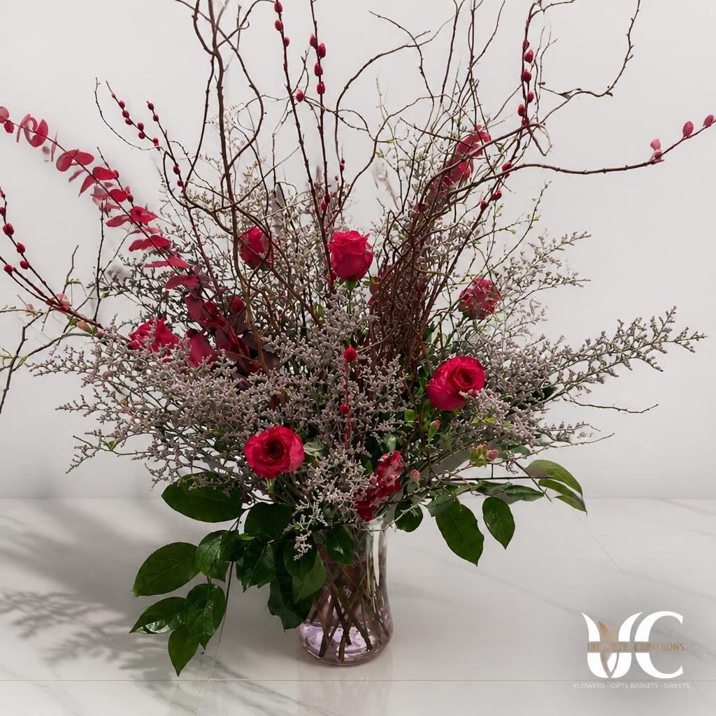 Tall clear glass vase arrangement with red roses, twisting bare branches, red berry-like accents, pale airy filler flowers, and glossy green leaves displayed on a sunlit tiled floor.