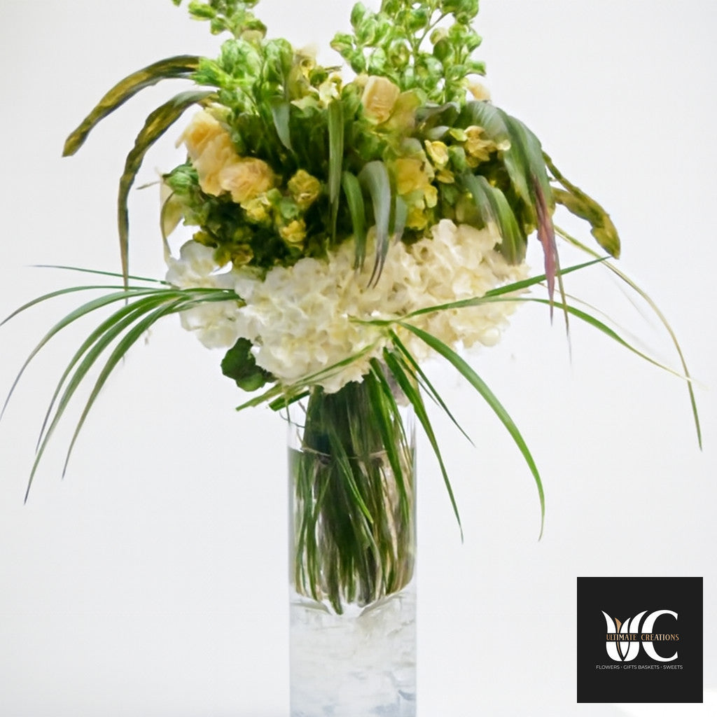 Tall glass vase arrangement with cream flowers, green blooms, and long arching foliage.