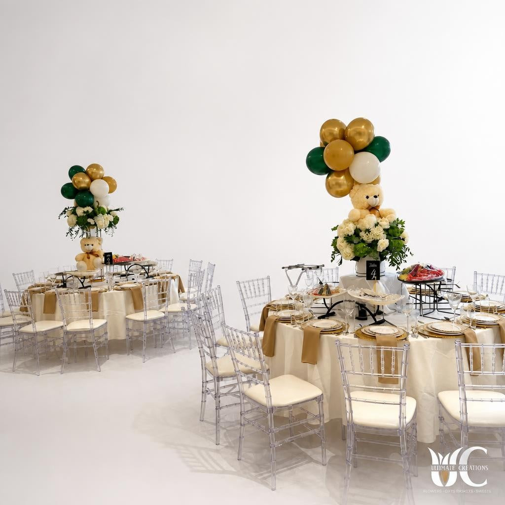 Bright event room with round tables, clear chairs, gold and green balloon clusters, floral accents, and teddy bear centerpieces.