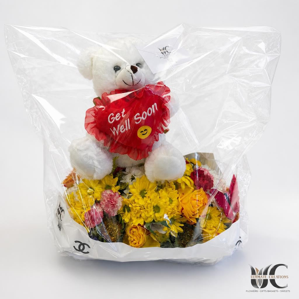 Cellophane-wrapped floral gift arrangement with a white teddy bear, red Get Well Soon heart, yellow flowers, orange and yellow roses, and pink accents.