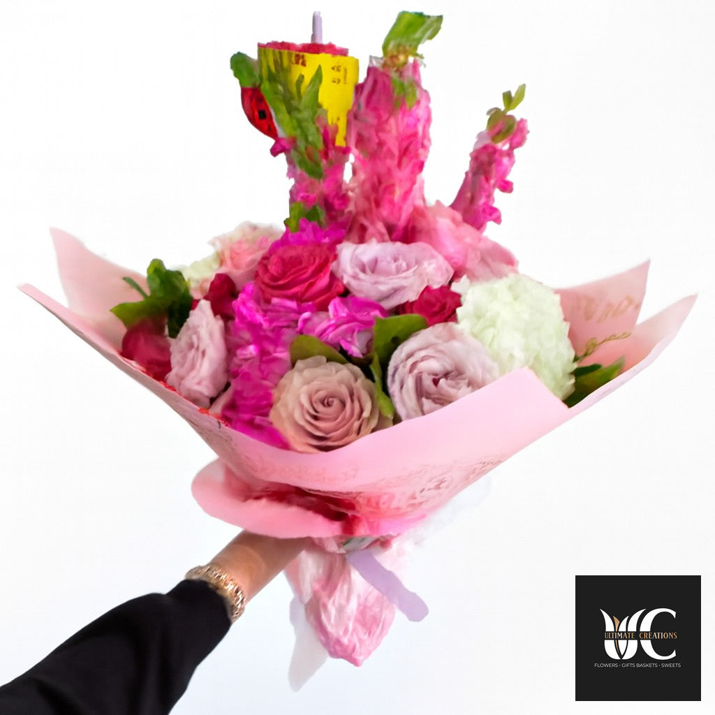 Hand-held pink bouquet with roses, blush flowers, and a decorative sweet-treat topper wrapped in pink paper.