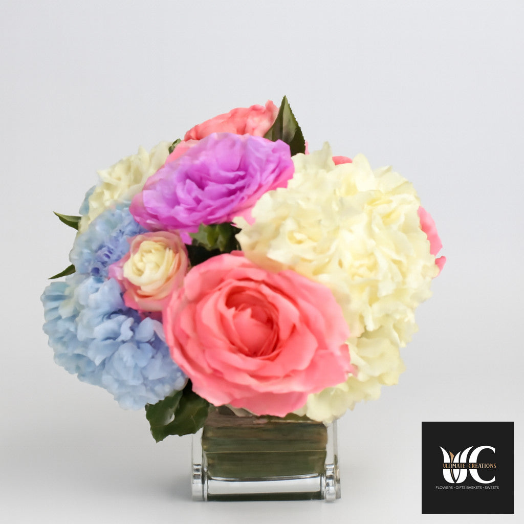 Pastel mixed flower arrangement in a clear cube vase with pink, lavender, cream, and light blue blooms.