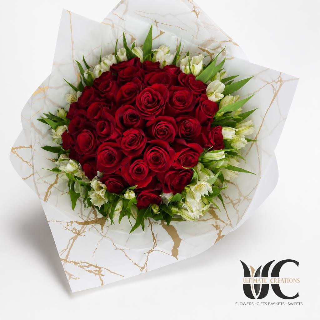 Round bouquet of red roses with white accent flowers and green foliage wrapped in white marble-pattern paper.