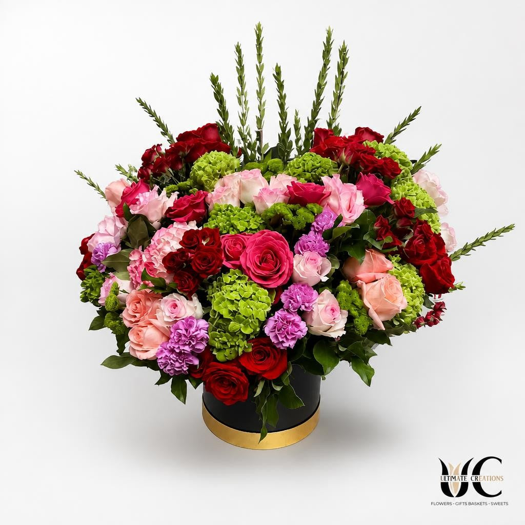 Large rounded floral arrangement with red and pink roses, green hydrangea, lavender flowers, peach blooms, and greenery in a black-and-gold hatbox.