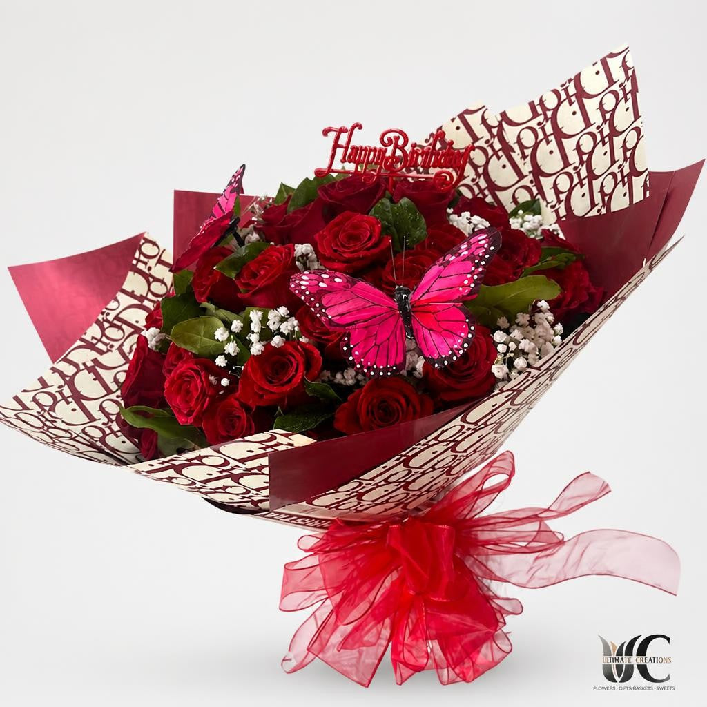 Red rose bouquet wrapped in patterned paper with white baby’s breath, green leaves, a large pink butterfly, red bow, and “Happy Birthday” topper.