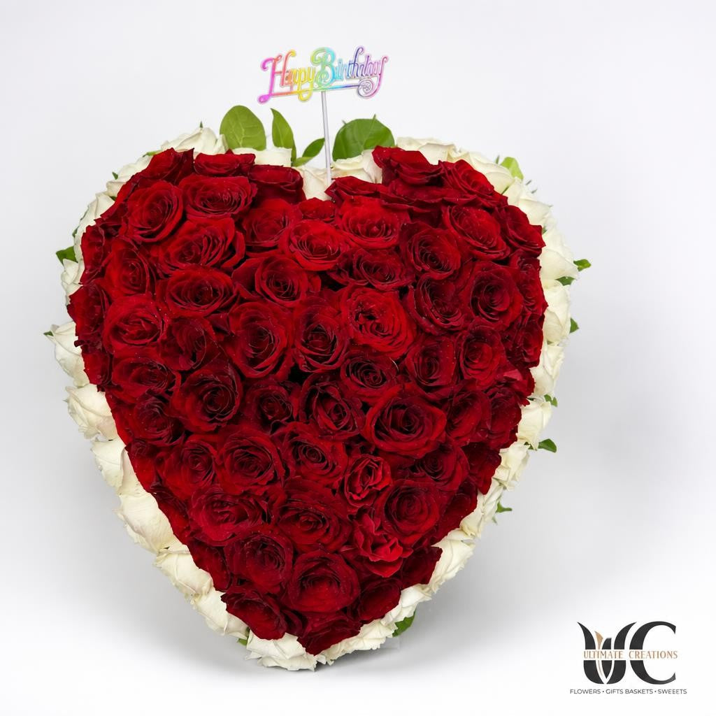 Heart-shaped floral arrangement densely covered with red roses, bordered by white roses, with a colorful birthday topper.