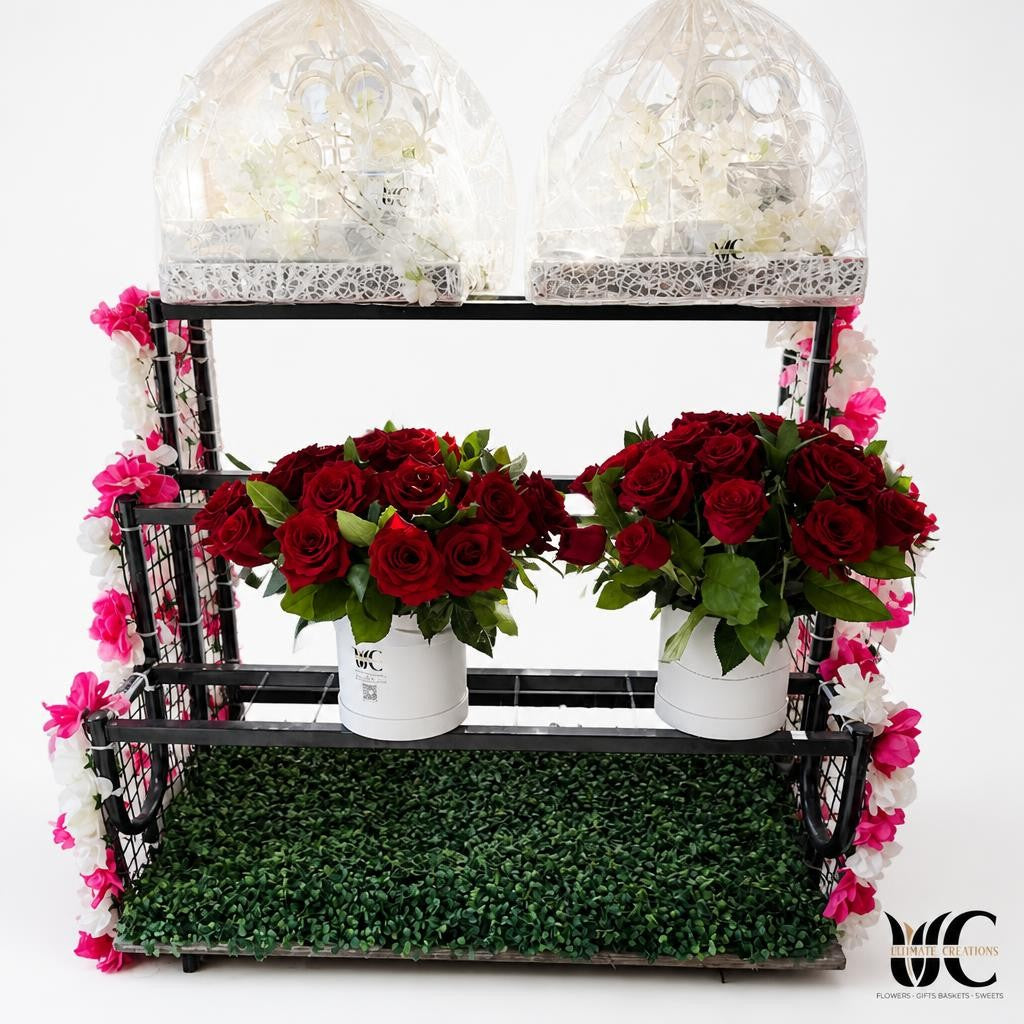 Two matching white containers holding rounded arrangements of deep red roses and green foliage on a display stand.