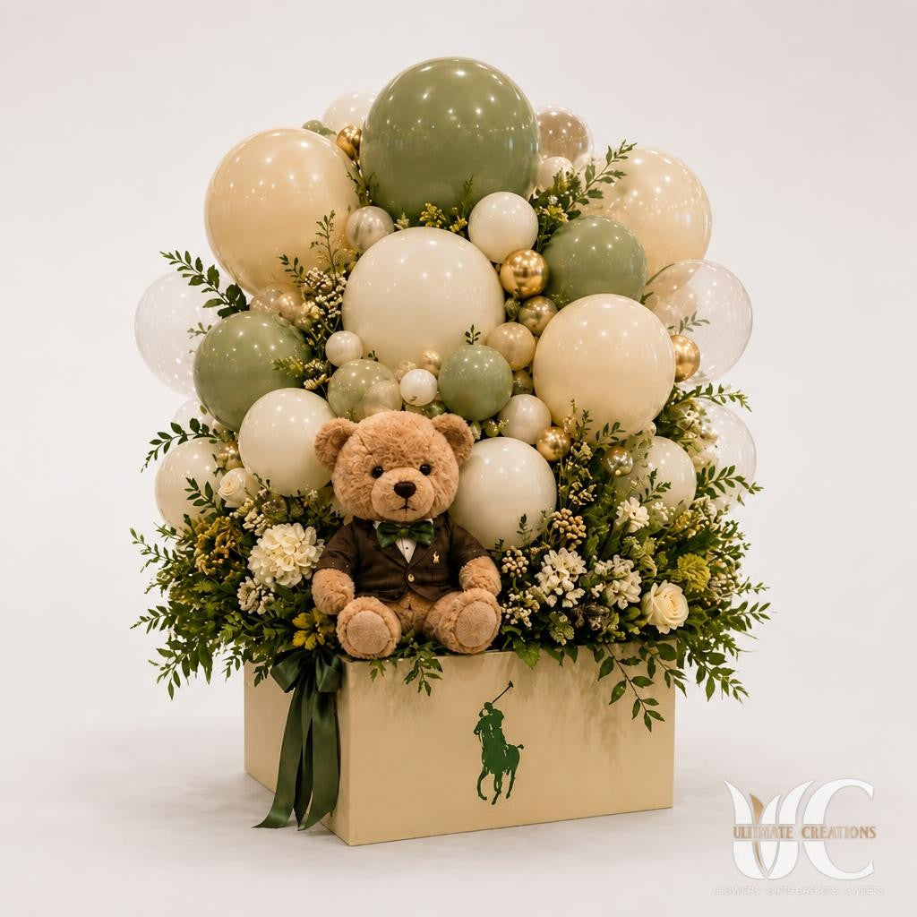 Large sage green, ivory, white, and gold balloon arrangement with flowers, greenery, and a teddy bear in a square presentation box on a formal dining table.