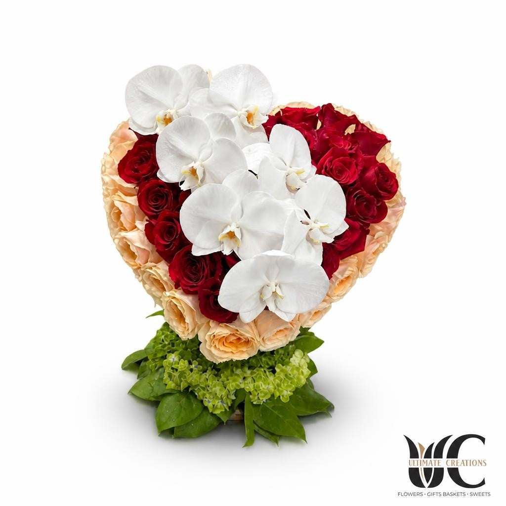 Heart-shaped floral arrangement with white orchids, red and blush roses, green hydrangea, and leafy foliage on a florist worktable.