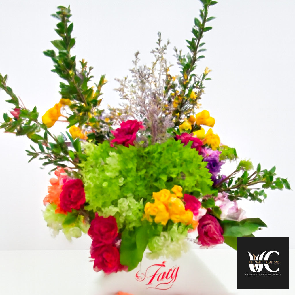 Colorful mixed flower vase arrangement with tall greenery, pink, yellow, orange, and purple blooms.