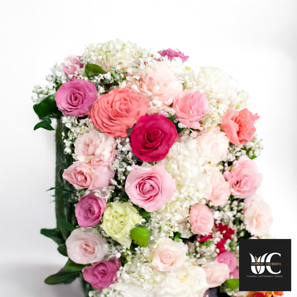 Cascade-style floral arrangement with pink, blush, coral, and white roses and small white filler flowers against a light background.