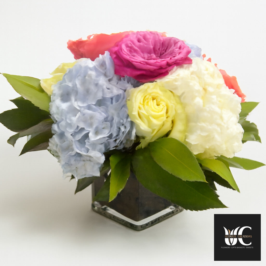 Colorful mixed flower arrangement in a clear square glass vase with pink, yellow, white, blue, and coral blooms and green leaves.