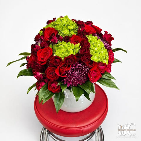 Lush red and burgundy flower arrangement with green blooms and purple accent in a white vase