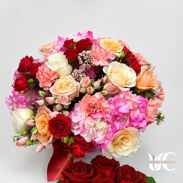 Lush hand-tied bouquet of red, pink, peach, cream, and white flowers with greenery and red ribbon.