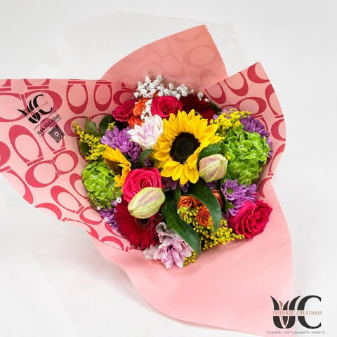 Colorful hand-tied bouquet in pink wrapping with a sunflower, roses, purple blooms, green flowers, and foliage