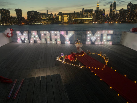 Full Adam and Gabriella engagement proposal setup at the Long Island City waterfront in Queens with illuminated letters, candles, red carpet and photos.