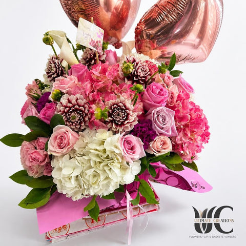 Lush pink and lavender flower bouquet with white hydrangea, greenery, pink wrapping, and rose-colored balloons in the background.