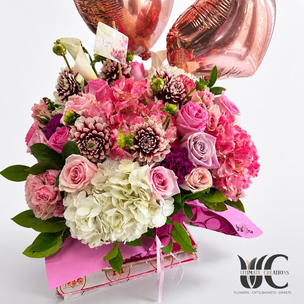 Lush pink and lavender flower bouquet with white hydrangea, greenery, pink wrapping, and rose-colored balloons in the background.