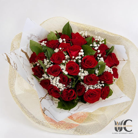 Top view of a rounded bouquet of red roses with white filler flowers and green leaves in white and metallic-textured wrapping.