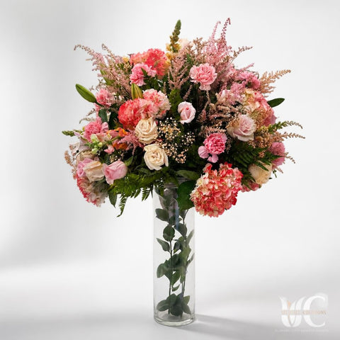 Tall glass vase arrangement with abundant blush, pink, cream, mauve, and green flowers