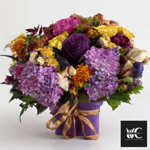 Purple flower box arrangement with mixed purple, lavender, yellow, and cream blooms tied with a gold ribbon.