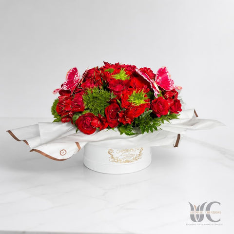 Low rounded arrangement of red roses, green accents, and pink butterfly decorations in a white presentation box on a counter.
