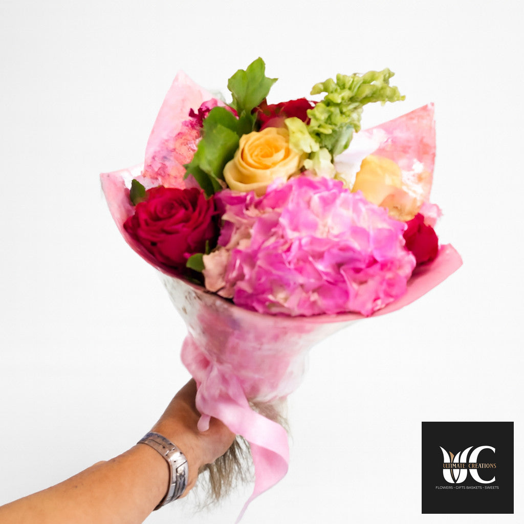 Hand-tied bouquet with pink, red, yellow, and green flowers wrapped in pink paper.