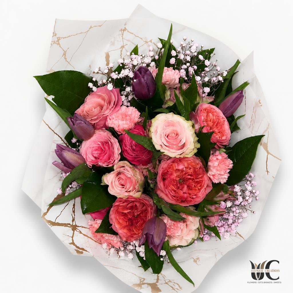 Hand-tied bouquet with pink, blush, coral, and lavender flowers wrapped in patterned white paper.