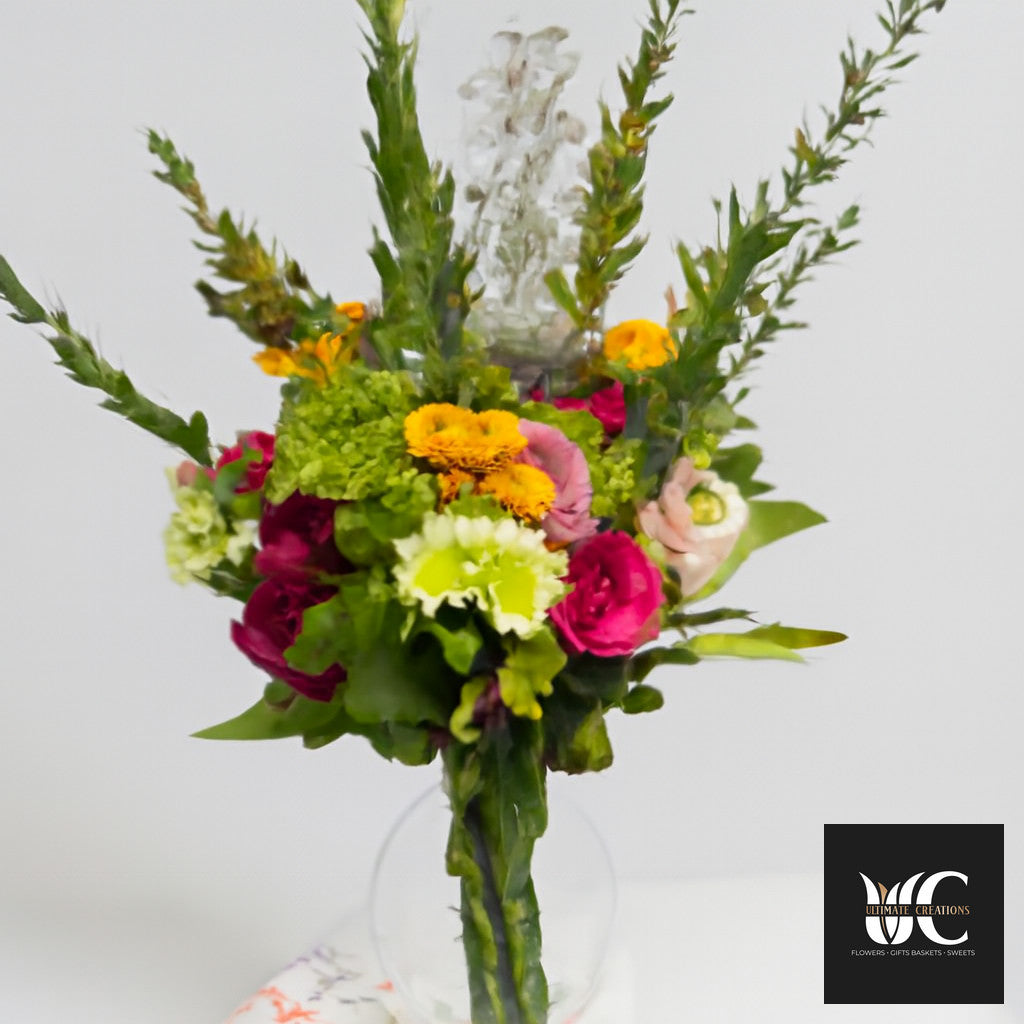 Tall mixed flower arrangement in a clear vase with green foliage, magenta and yellow flowers, and pink accents.