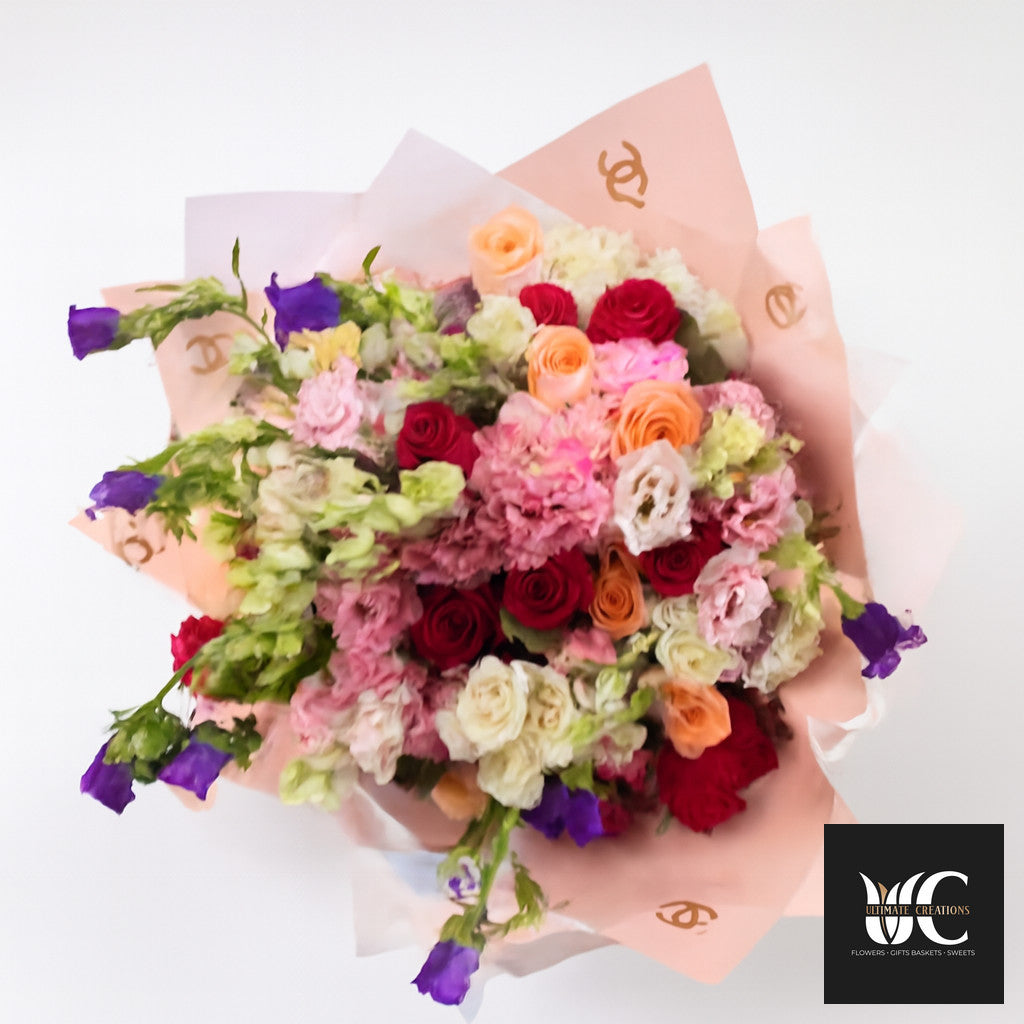 Large mixed bouquet with pink, red, peach, cream, and purple flowers wrapped in pastel paper.