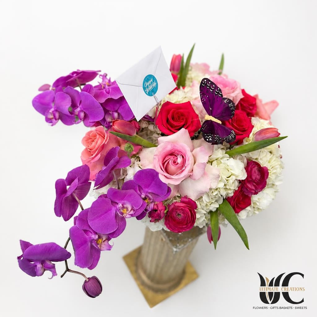 Lush floral arrangement in a clear glass vase with cascading purple orchids, pink and red roses, white clustered blooms, green leaves, and a decorative butterfly accent.