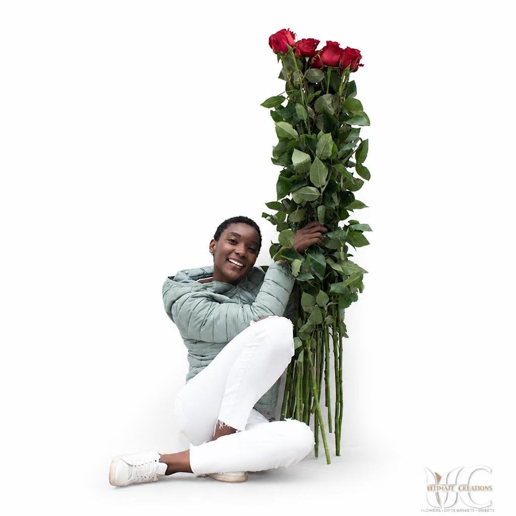 Tall bouquet of red roses with long green stems and foliage against a white wall.