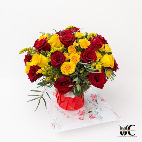 Rounded floral arrangement of red and yellow roses with green foliage and small yellow filler flowers in a red container.