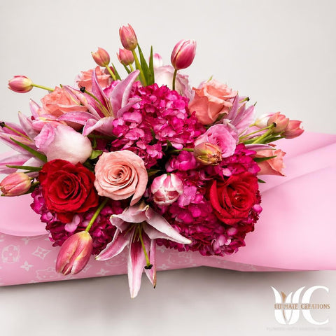 Lush bouquet featuring pink hydrangeas, blush and burgundy roses, pink lilies, and tulips with green stems and leaves.