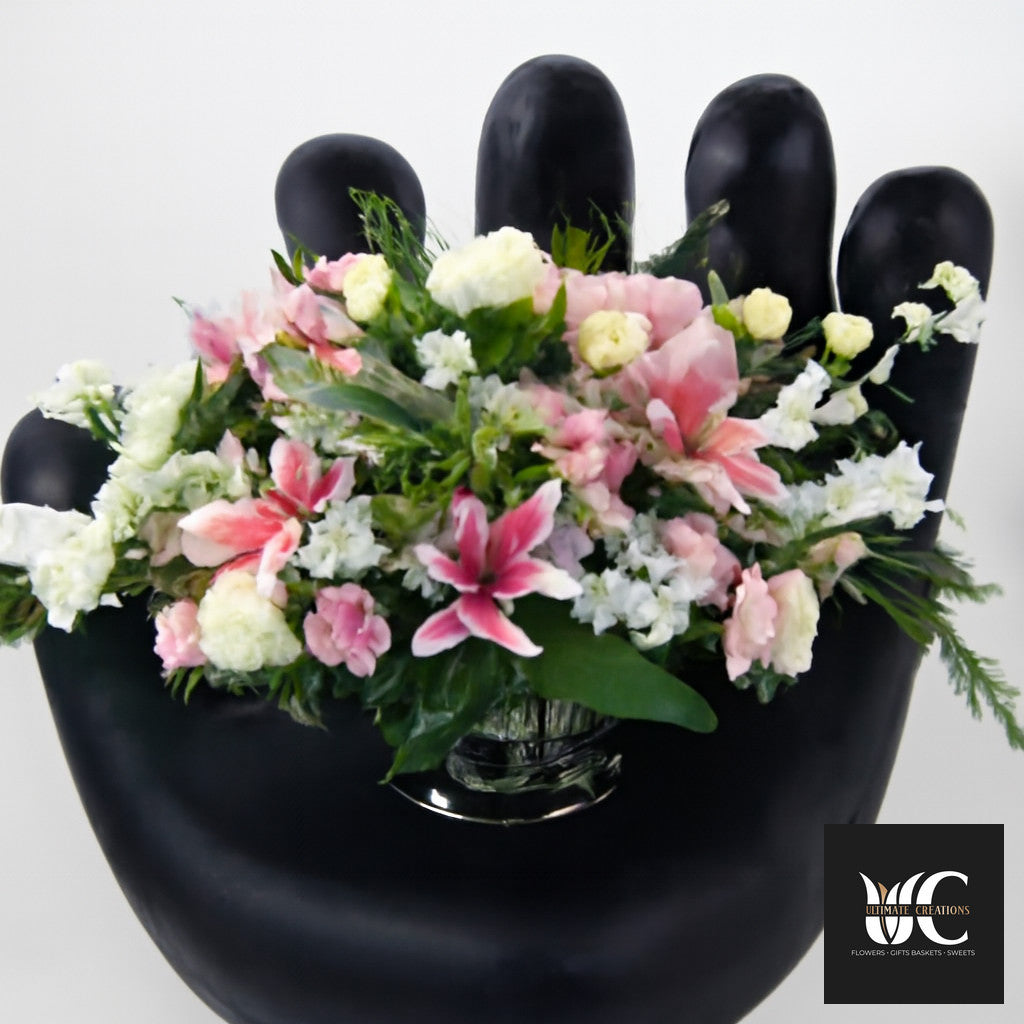 Pink and white floral arrangement in a black hand-shaped vase with greenery.