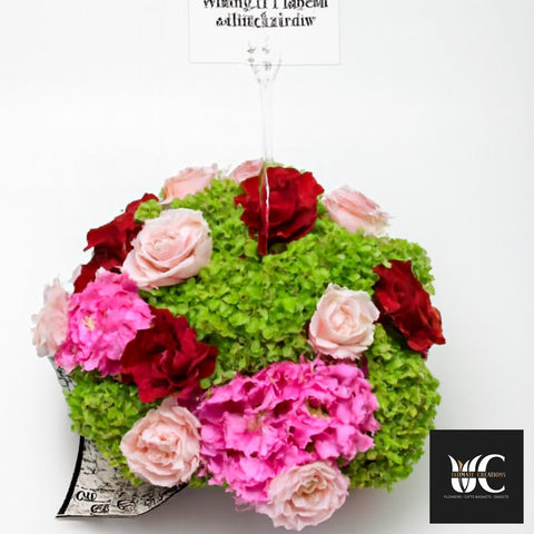 Round flower box arrangement with red, blush, and pink flowers on a green hydrangea base.