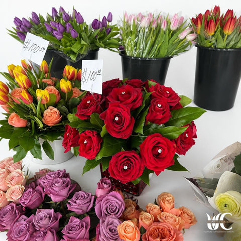 Bouquet of red roses with glossy green leaves and small white decorative accents, displayed among purple, peach, yellow, pink, and white flowers on florist shelving.