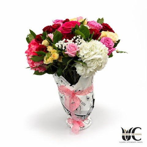 Lush wrapped bouquet of pink, red, cream, lavender, and white flowers with greenery and a pink ribbon