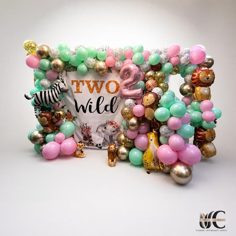 Indoor balloon display with mint, pink, white, gold, bronze, and charcoal balloons surrounding a “Two Wild” backdrop, oversized number two, animal-shaped balloons, and jungle-themed accents beneath a chandelier.