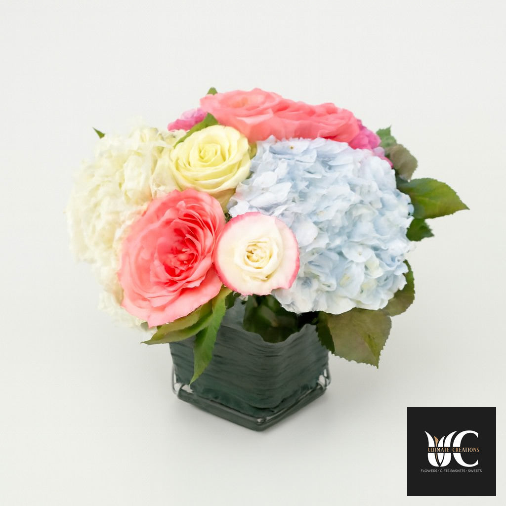 Pastel roses and hydrangea arranged in a square glass vase with green leaves.