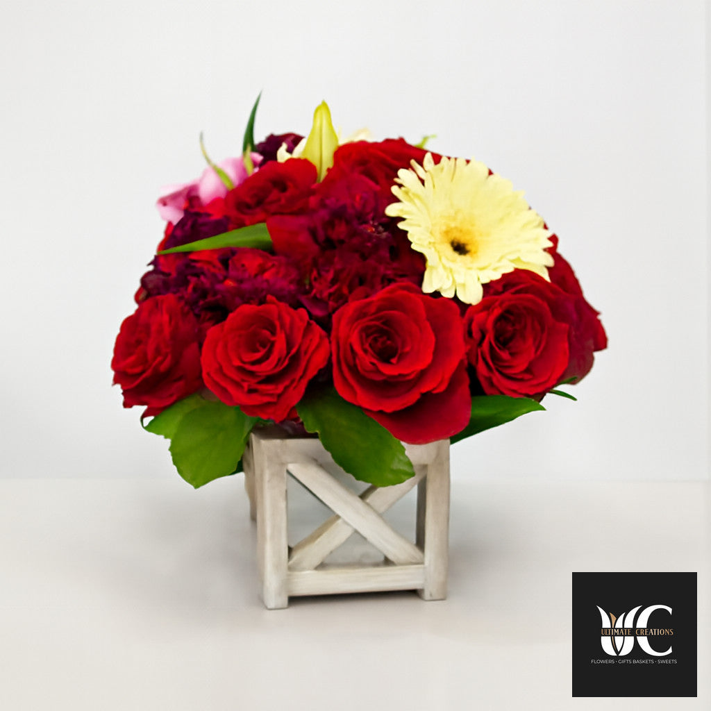 Compact floral centerpiece with red roses, a cream daisy, pink accents, and greenery in a wooden cube vase.